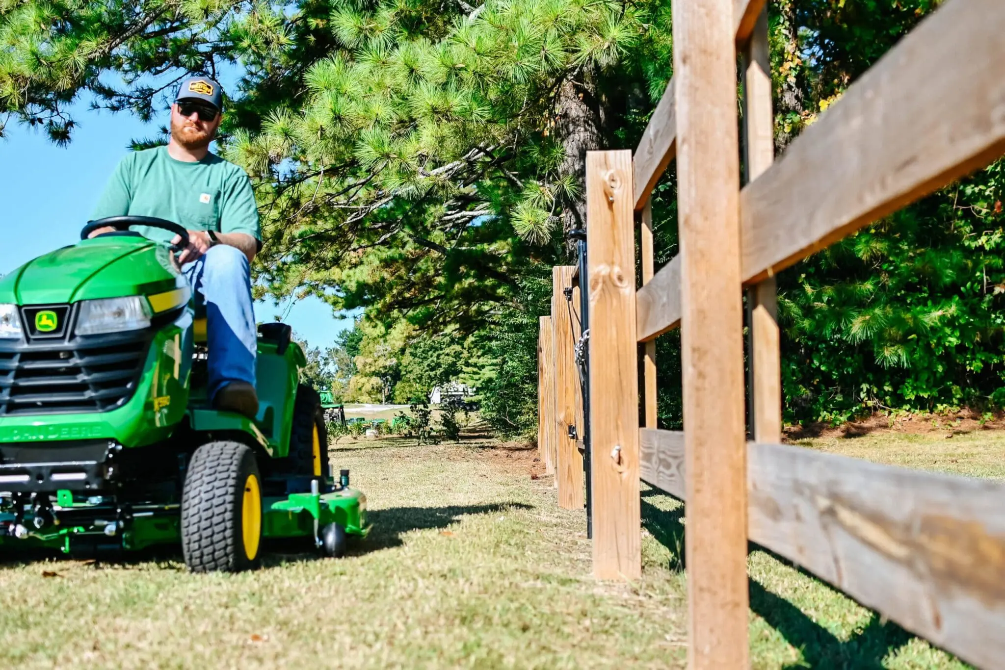 A man drives a John Deere gas riding lawn tractor near a fence.