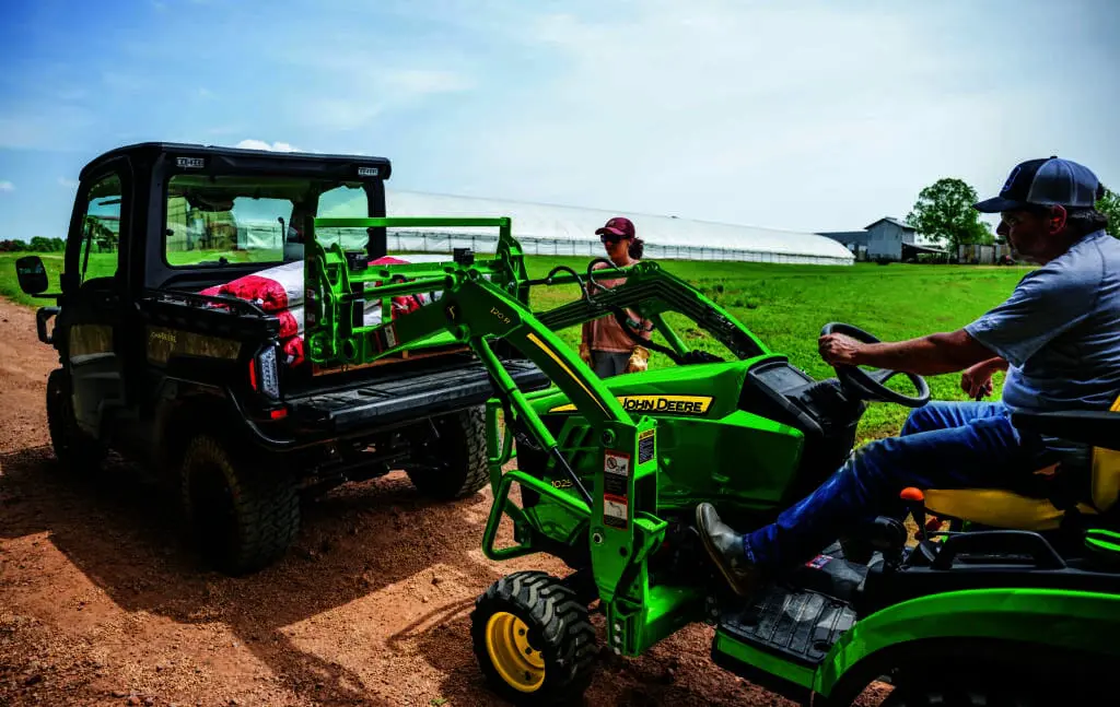 A man loads a pallet into the back of a John Deere Gator using a compact utility tractor 