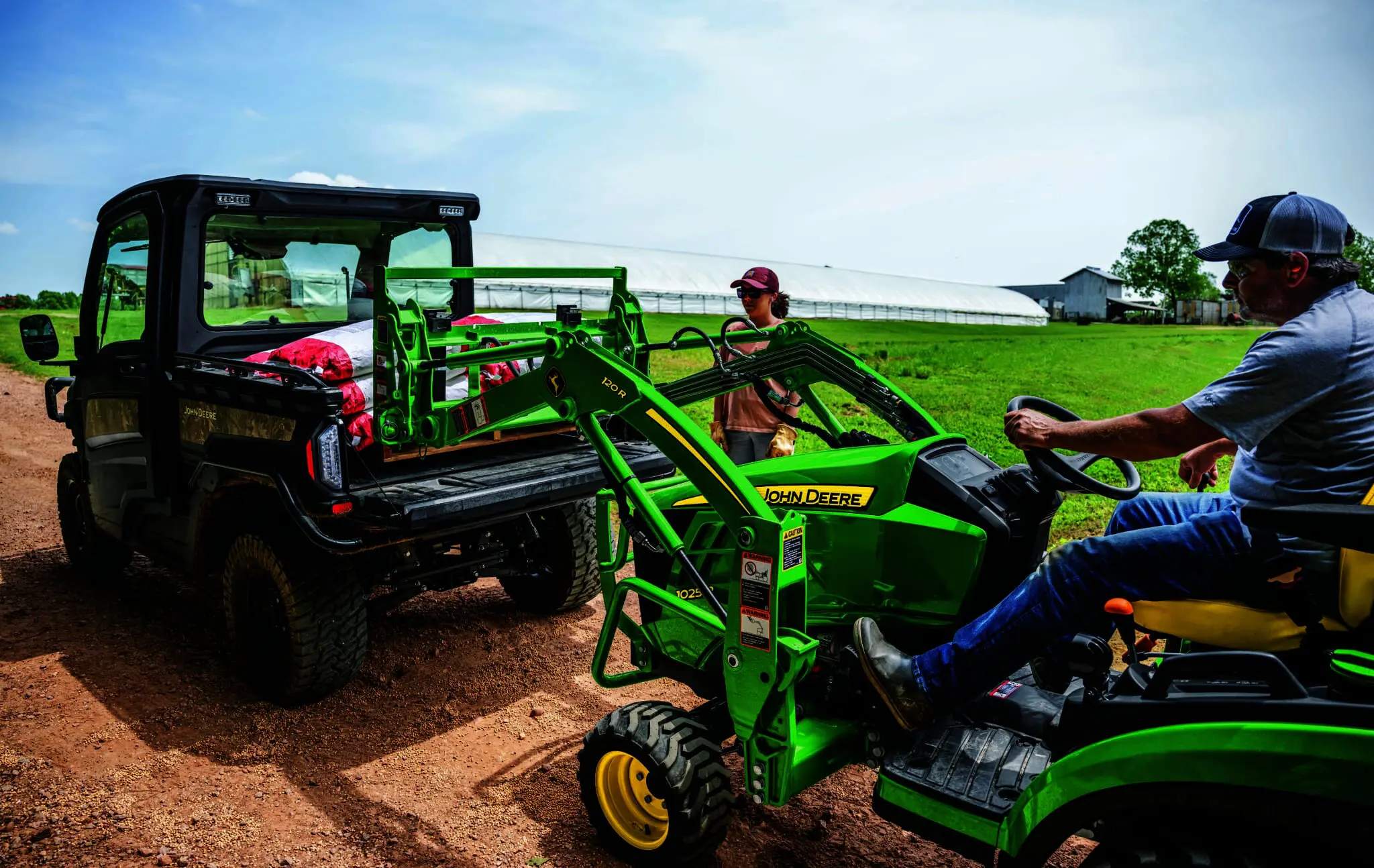 A man loads a pallet into the back of a John Deere Gator using a compact utility tractor