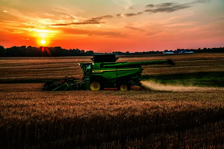 A John Deere S7 combine harvests wheat in a farm field. The sun sets in the background.