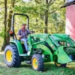 A man carries a load of plants in a Deere 3R tractor loader next to a red barn