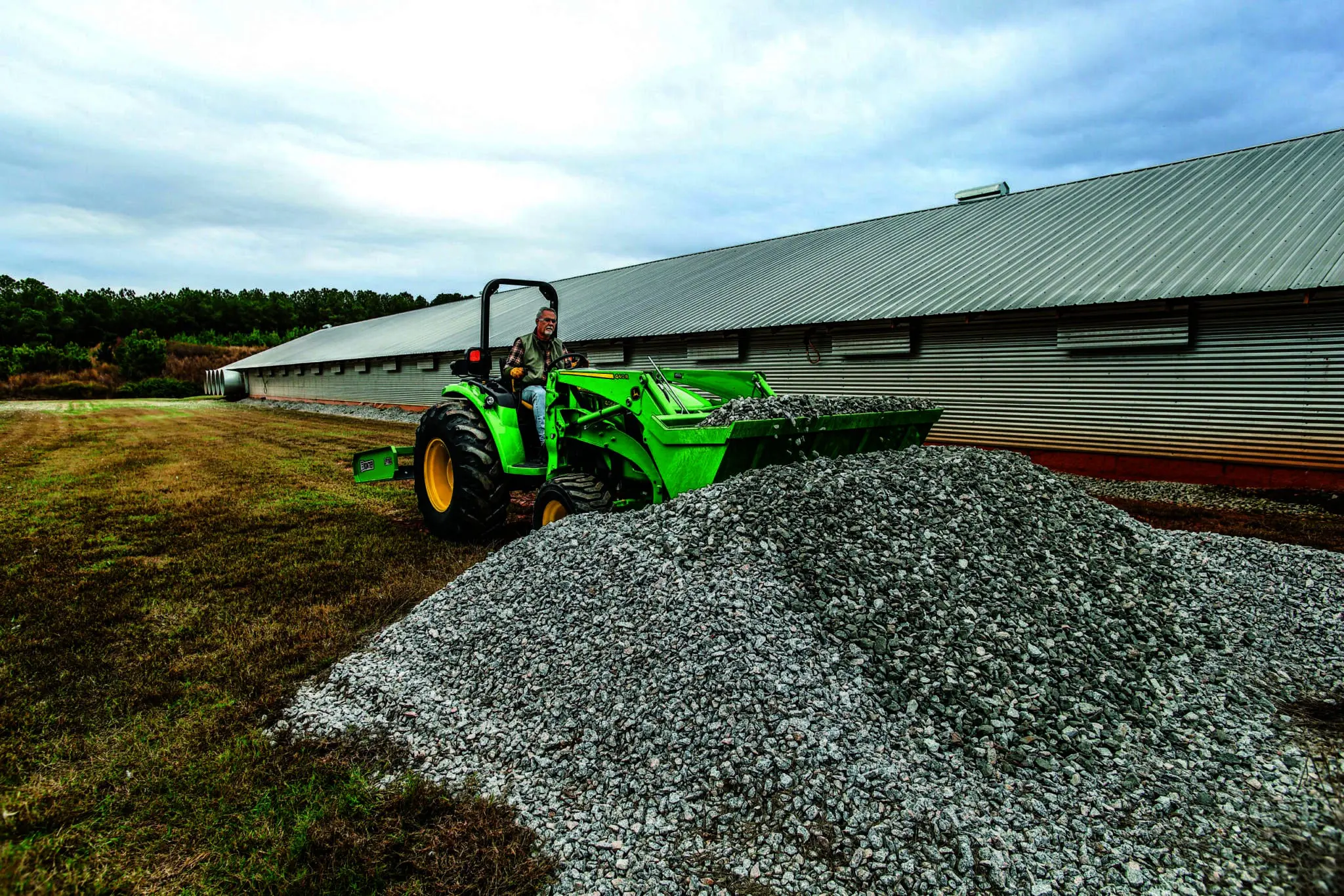 A person moves gravel next to a barn with a Deere 4M tractor loader