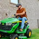 A man mows next to a brick house with a Deere S100 lawn tractor