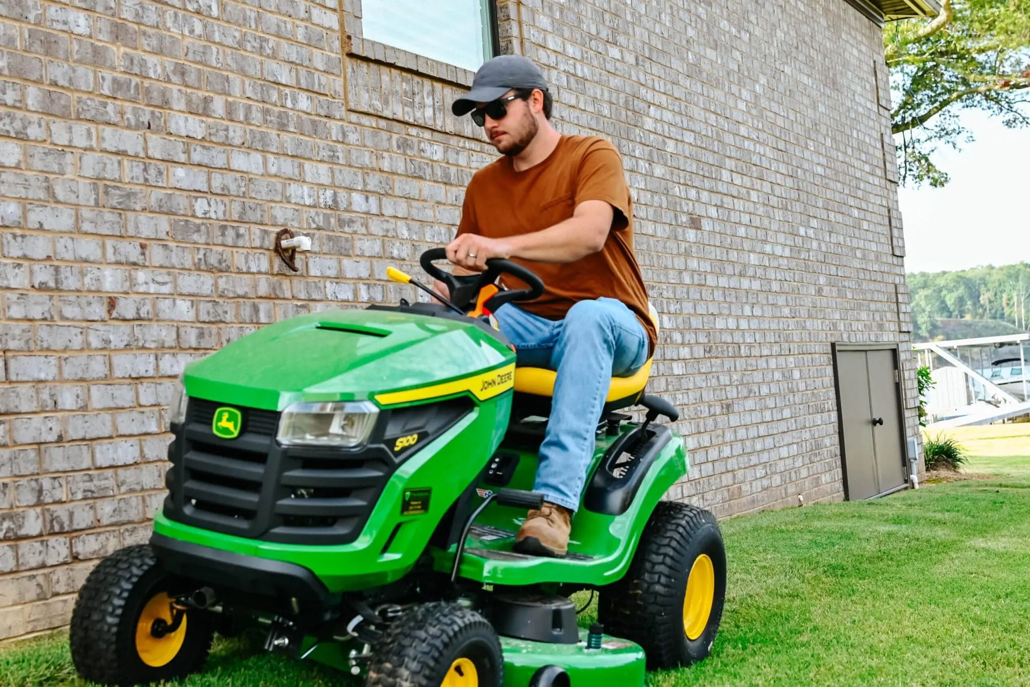 A man mows next to a brick house with a Deere S100 lawn tractor