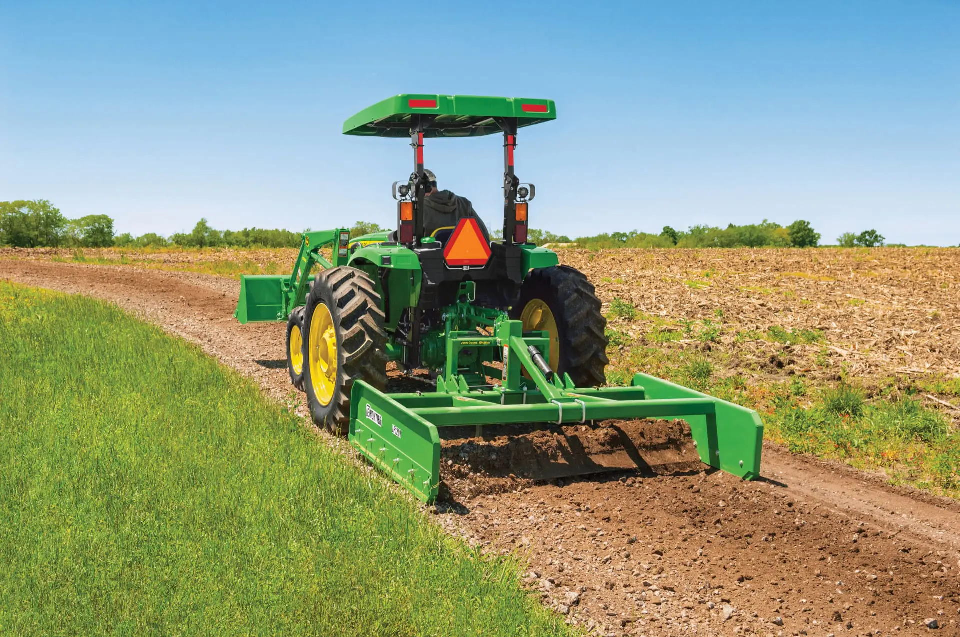 A person uses John Deere tractor and a land plane on a dirt road