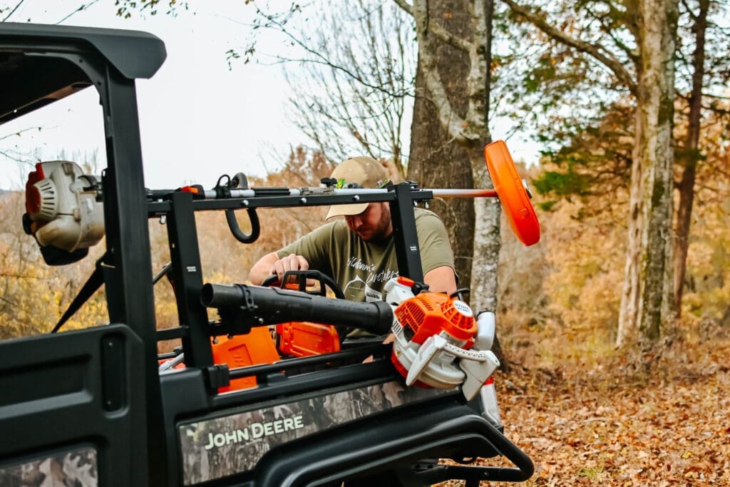 A person loads STIHL power tools including a weed eater, chain saw and leaf blower into the back of a John Deere Gator utility vehicle