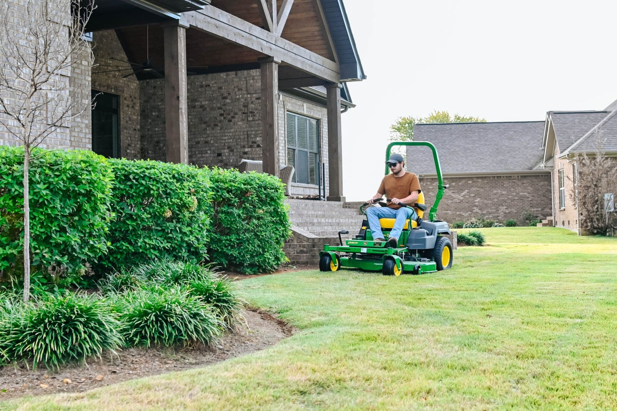 A person uses a John Deere zero turn mower to mow along bushes by a house