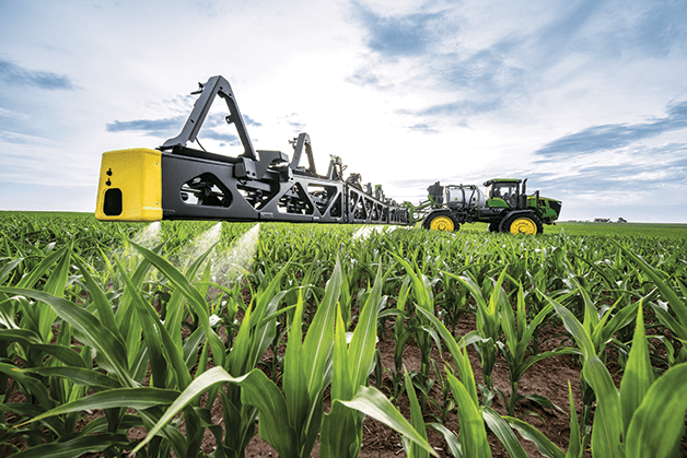 A John Deere sprayer with See and Spray precision technology applies chemicals in a corn field.