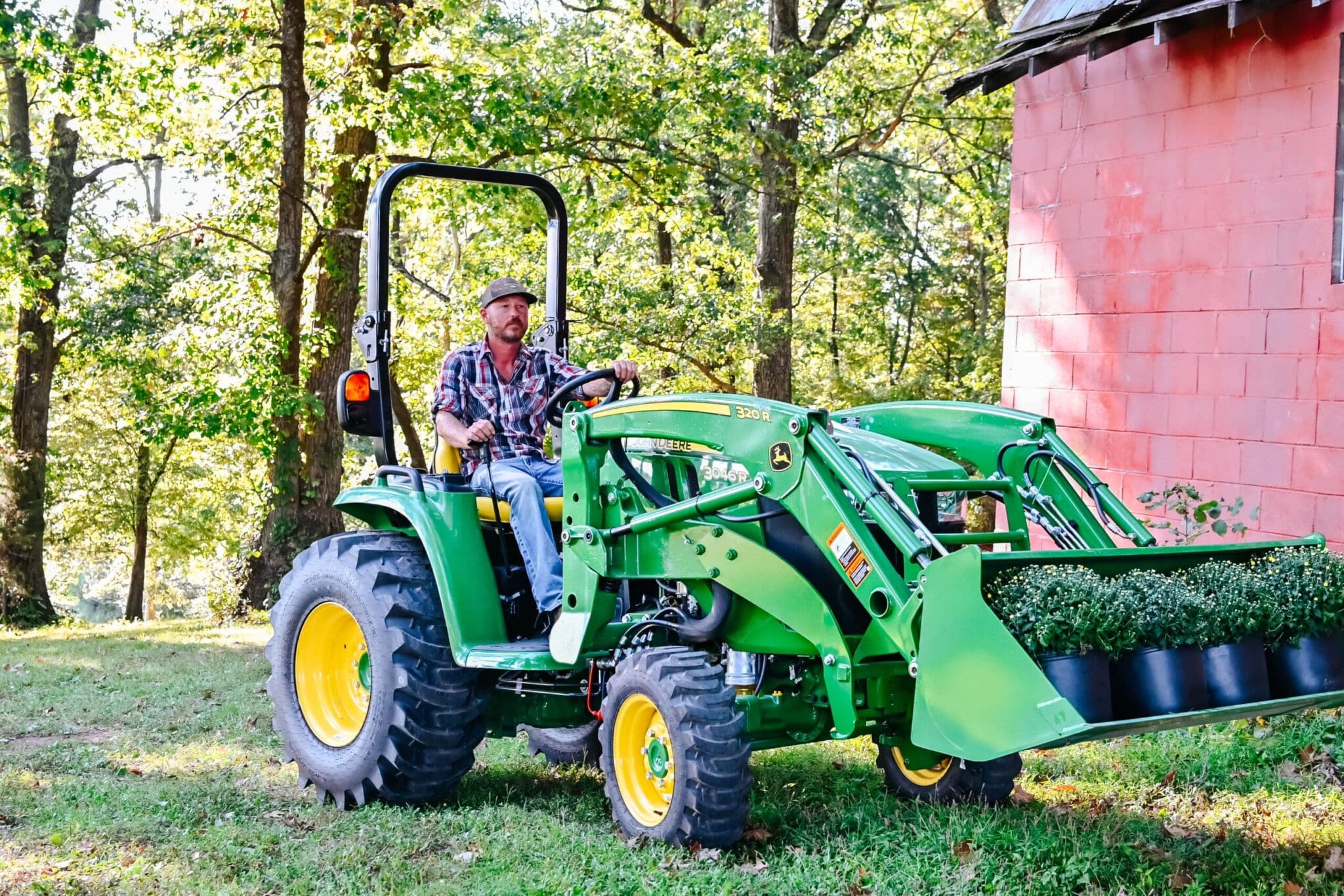 A man carries a load of plants in a Deere 3R tractor loader next to a red barn