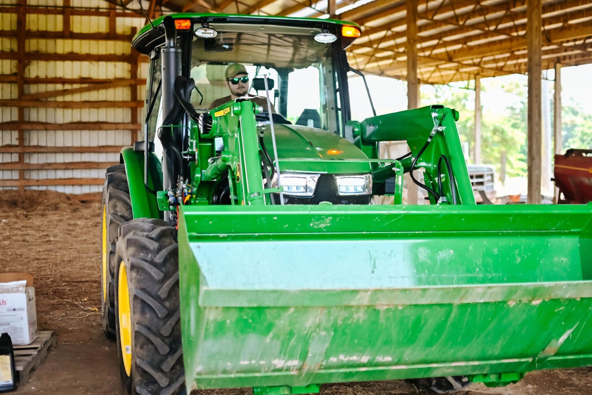 A man moves a cab 5E John Deere tractor with loader inside a barn
