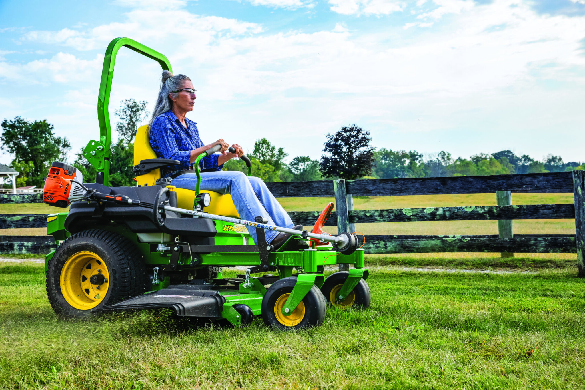 A woman mows along a fence with a Deere Z735M zero turn mower
