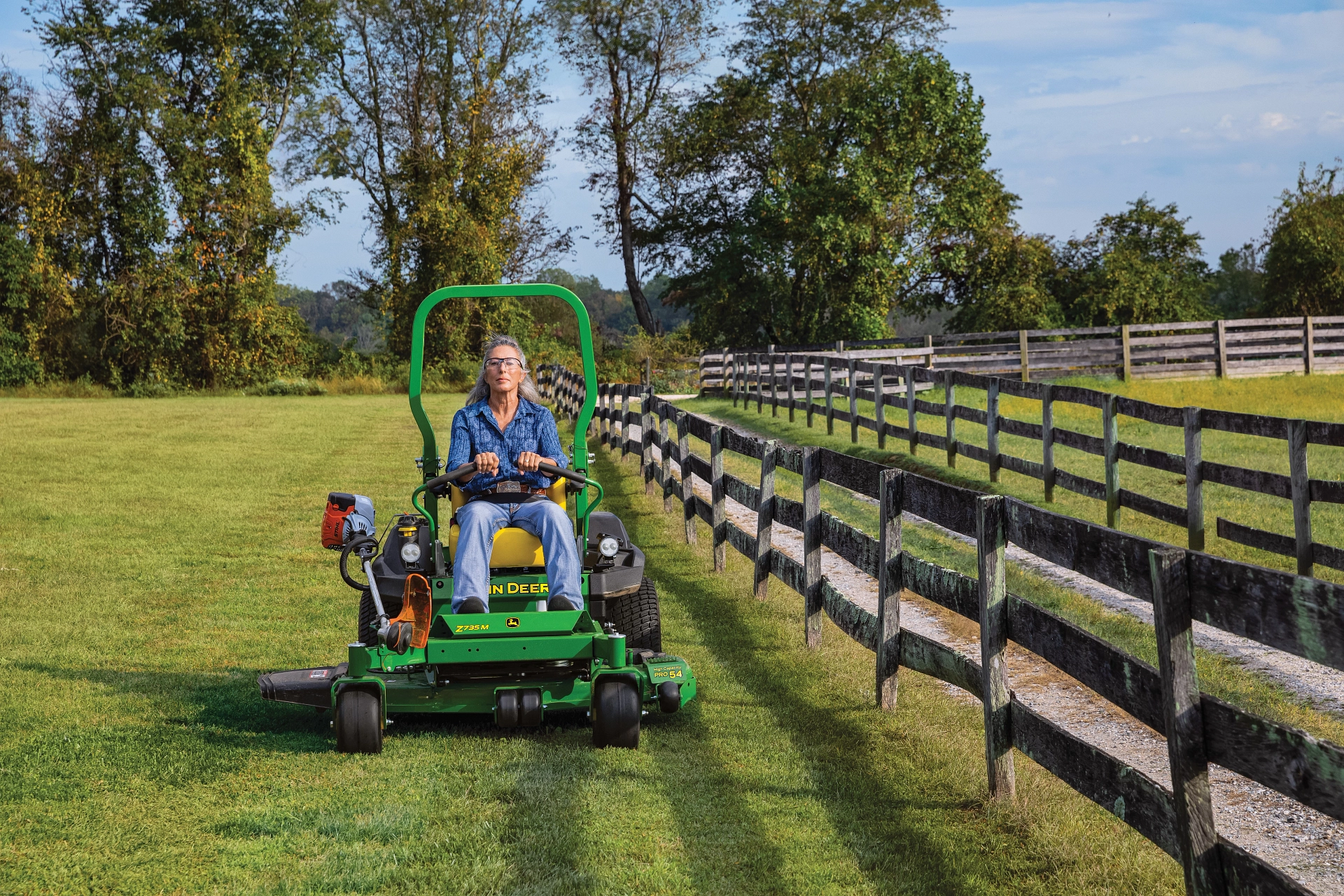 Woman mows lawn with John Deere Z735M ZTrak zero turn lawn mower next to split rail fence